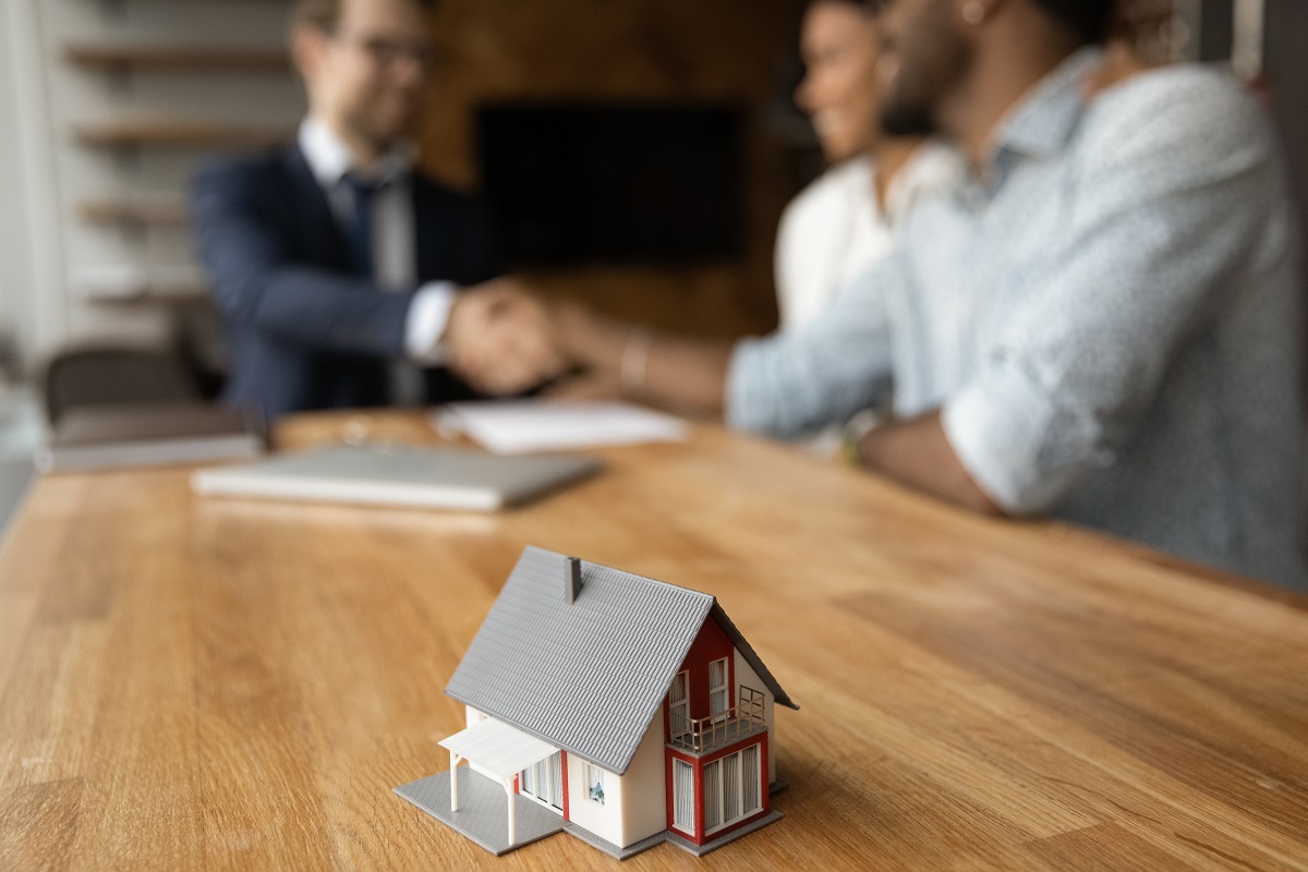 Close up focus on little mansion house model on table with blurred joyful african american clients shaking hands with professional realtor on background, real estate services, mortgage payment concept